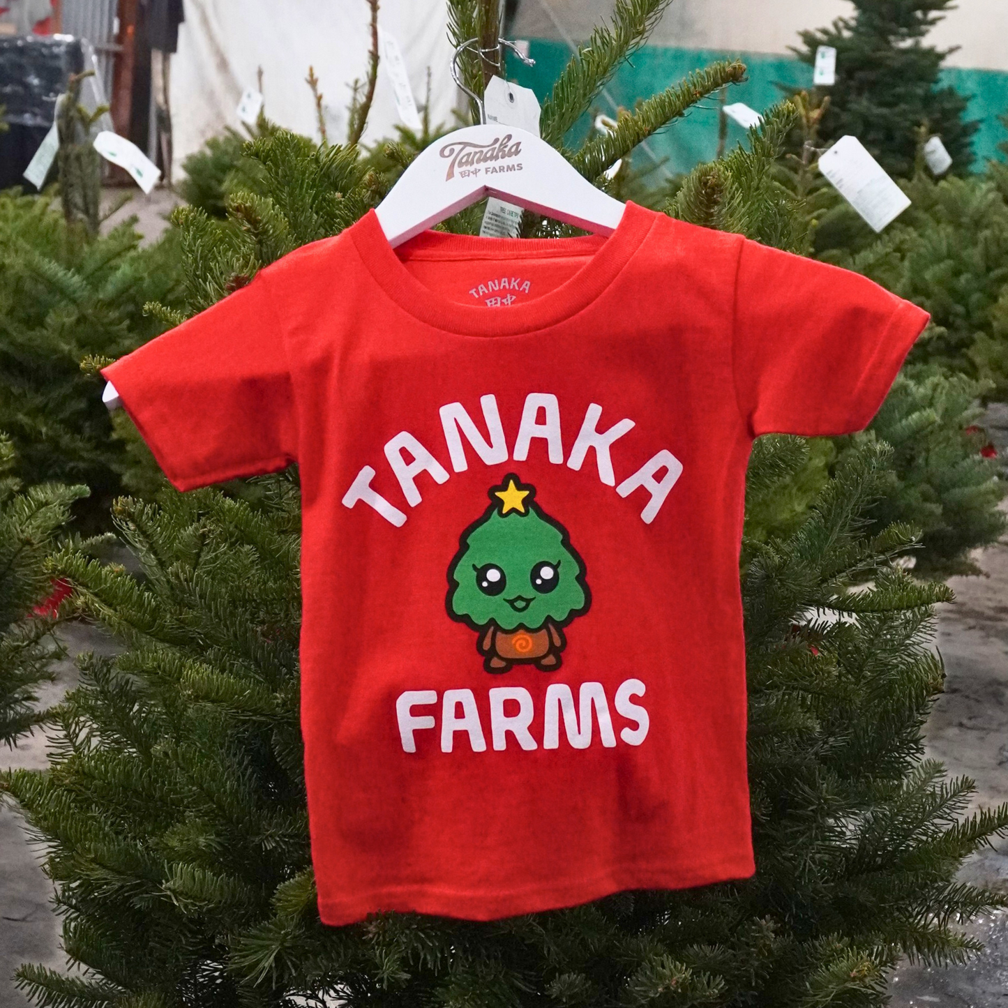 Red t-shirt with 'Tanaka Farms' text and logo on a hanger amidst Christmas trees.